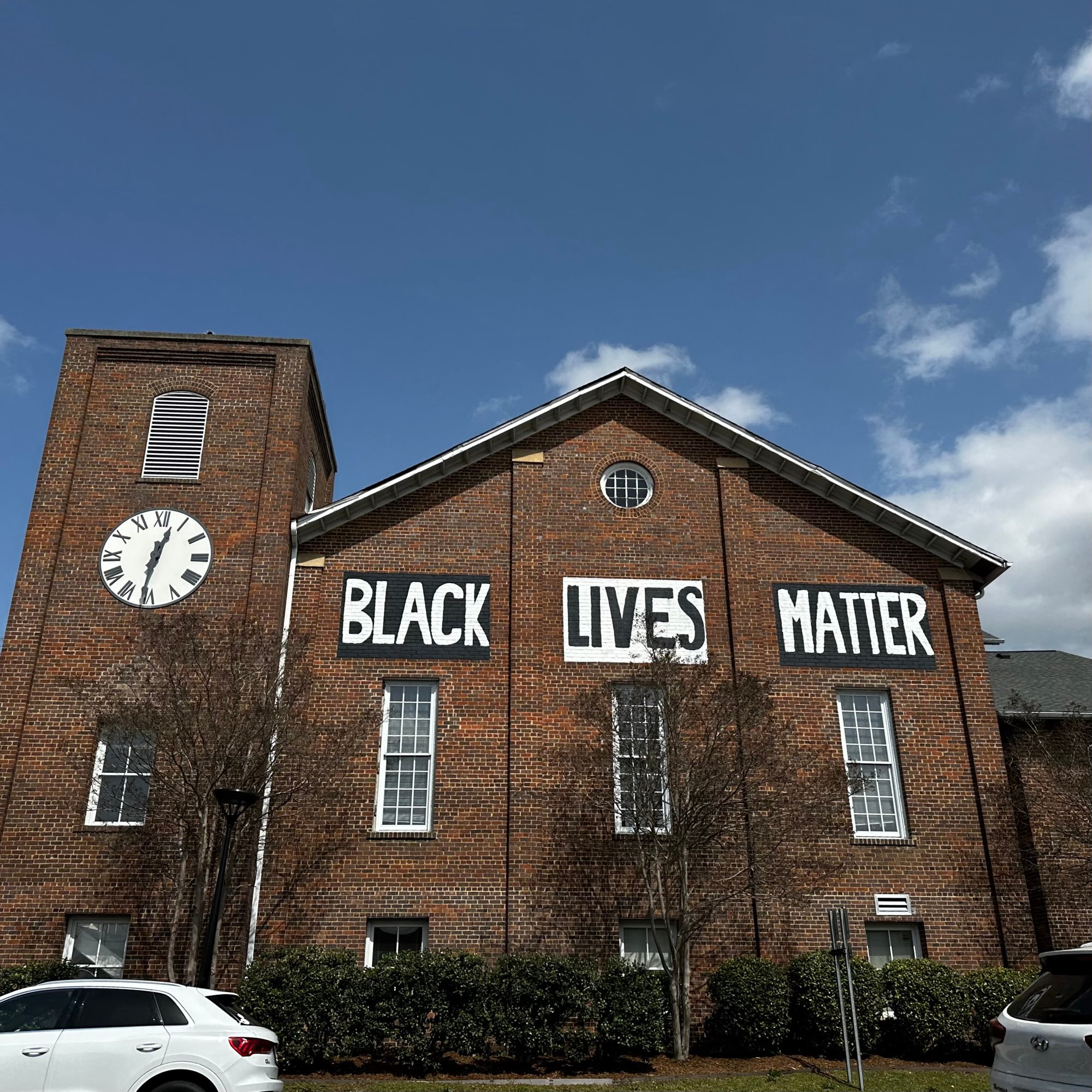 A picture from street level looking north. A red brick building fills the bottom two thirds of the frame, with partly cloudy skies above. The building has "Black Lives Matter" painted by hand in black and white.
